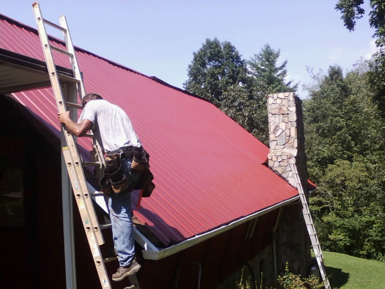 Roofer on a ladder beside a red metal roof and stone chimney — Soffit & Fascia Repair by Roofing Brothers in Glennville